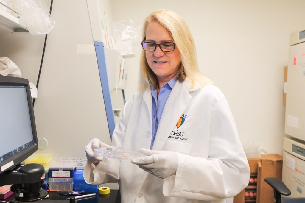 Researcher Rosalie is in a lab coat and gloves and holds a clear multi-well plate beside laboratory equipment in a biomedical research lab at the Robertson Life Sciences Building in Portland, Oregon.